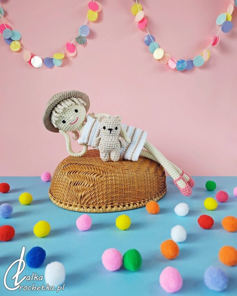 Personalizowana szydełkowa lalka w letnim kapeluszu i sukience w paski leży na wiklinowym koszyku obok małego misia przyjaciela, na tle różowo-niebieskiej scenografii z kolorowymi pomponami i girlandą