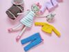 Personalizowana lalka w pasiastej sukience otoczona kompletem zdejmowanych ubranek: niebieskimi spodniami, żółtym i fioletowym sweterkiem, brązowym plecakiem, kapeluszem oraz miniaturowym misiem na różowym tle